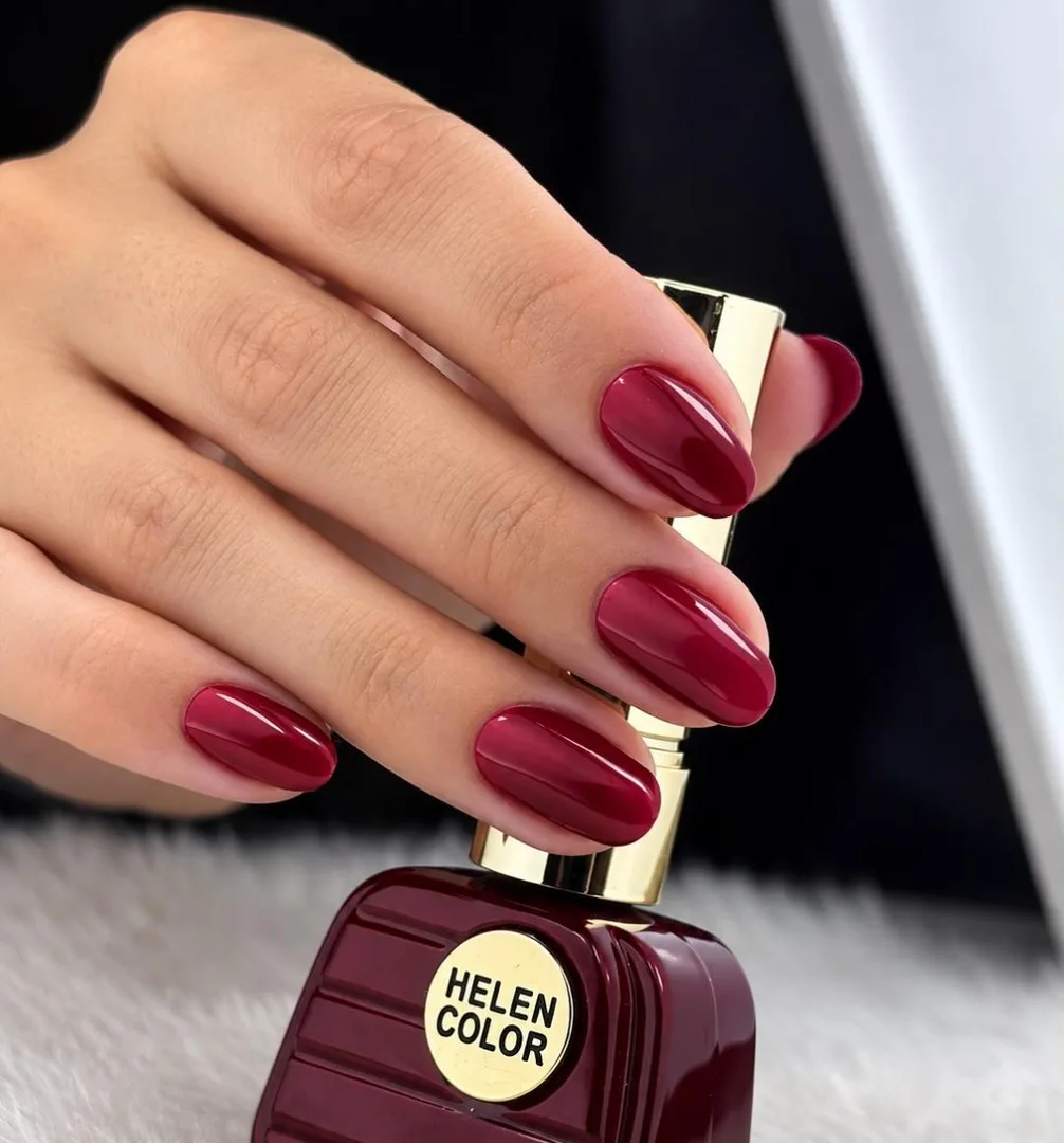 Mão com unhas amendoadas esmaltadas em bordô brilhante, segurando um vidro de esmalte da mesma cor